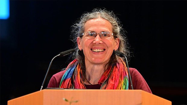 Eine Frau mit grauem, lockigem Haar und Brille steht hinter einem Holzpult und lächelt. Sie trägt einen bunten Schal und ein dunkelrotes Oberteil. Zwei Mikrofone sind vor ihr am Pult befestigt. Der Hintergrund ist dunkel und lenkt den Fokus auf die Person.