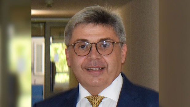Ein Mann mit grauem, seitlich gescheiteltem Haar trägt einen dunkelblauen Anzug, ein weißes Hemd und eine gelbe Krawatte mit feinem Muster. Der Hintergrund zeigt einen unscharfen Flur mit Türen und Fenstern.