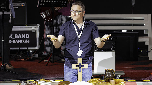 Das Bild zeigt einen Mann mittleren Alters mit kurzen, dunklen Haaren und einer Brille. Er trägt ein dunkles, ärmelloses Hemd und Jeans. Er steht vor einem hölzernen Kreuz und hält in jeder Hand ein kleines Brot. Im Hintergrund ist eine Bühne mit Musikinstrumenten zu sehen.