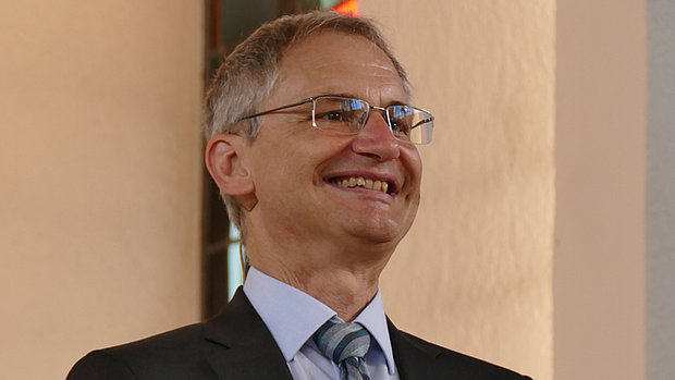 Ein Mann mit grauem, kurzem Haar und Brille trägt einen dunklen Anzug, ein hellblaues Hemd und eine gestreifte Krawatte. Er lächelt breit und blickt leicht zur Seite. Im Hintergrund ist eine helle Wand zu sehen.