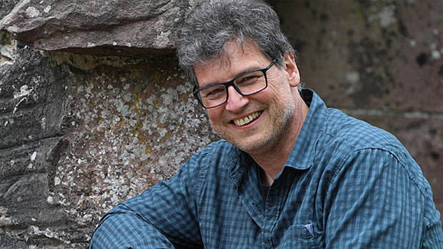 Porträt von Wilfried Röcker. Ein lächelnder Mann mittleren Alters mit grau melierten Haaren und einer Brille. Er trägt ein kariertes Hemd und sitzt vor einer Steinmauer.