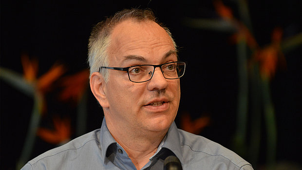 Portrait von Christof Voigt. Ein Mann mittleren Alters mit kurzen, grau melierten Haaren und einer Brille. Er trägt ein hellblaues Hemd und spricht in ein Mikrofon. Der Hintergrund ist unscharf und zeigt orangefarbene Blüten.