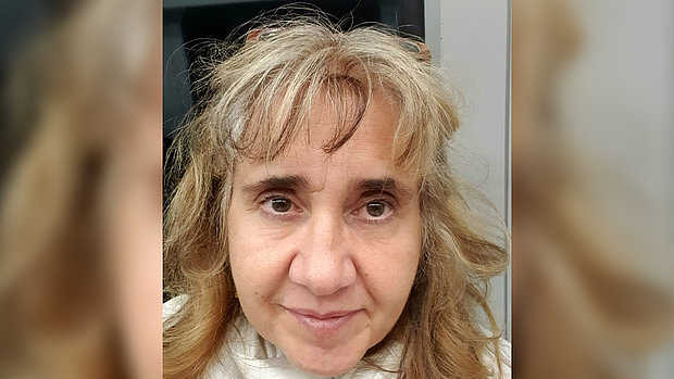 Porträt einer Frau mit schulterlangen, gewellten Haaren und neutralem Gesichtsausdruck vor unscharfem Hintergrund